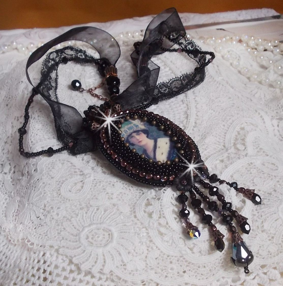 Pendentif Midnight Daisy avec un cabochon en bois repr&eacute;sentant une femme en chapeau Bleu, brod&eacute; avec des fleurs lucites, coupelles et facettes. Le tout est fini avec une dentelle Noire et un ruban