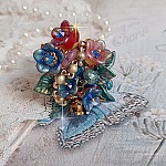 Bague Bel Air mont&eacute;e avec des fleurs lucites peintes &agrave; la main aux couleurs : Bleu, Rose, Rouge, Argent, Blanc avec feuilles ondul&eacute;es Vert Cuivr&eacute; &agrave; laquelle s&rsquo;ajoute perles Dor&eacute;es en Verre